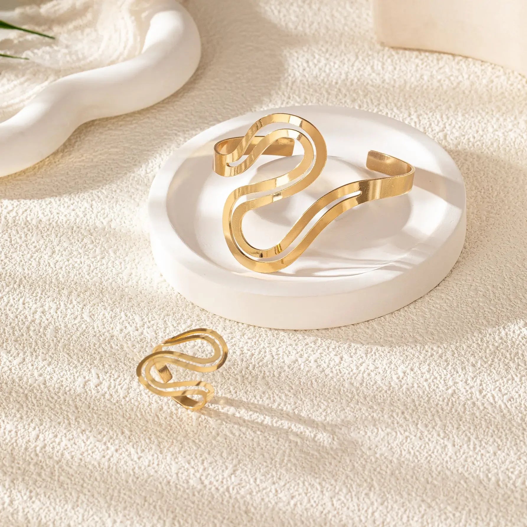 Gold wave snake-inspired bracelet and ring set displayed on a white ceramic dish against a soft background