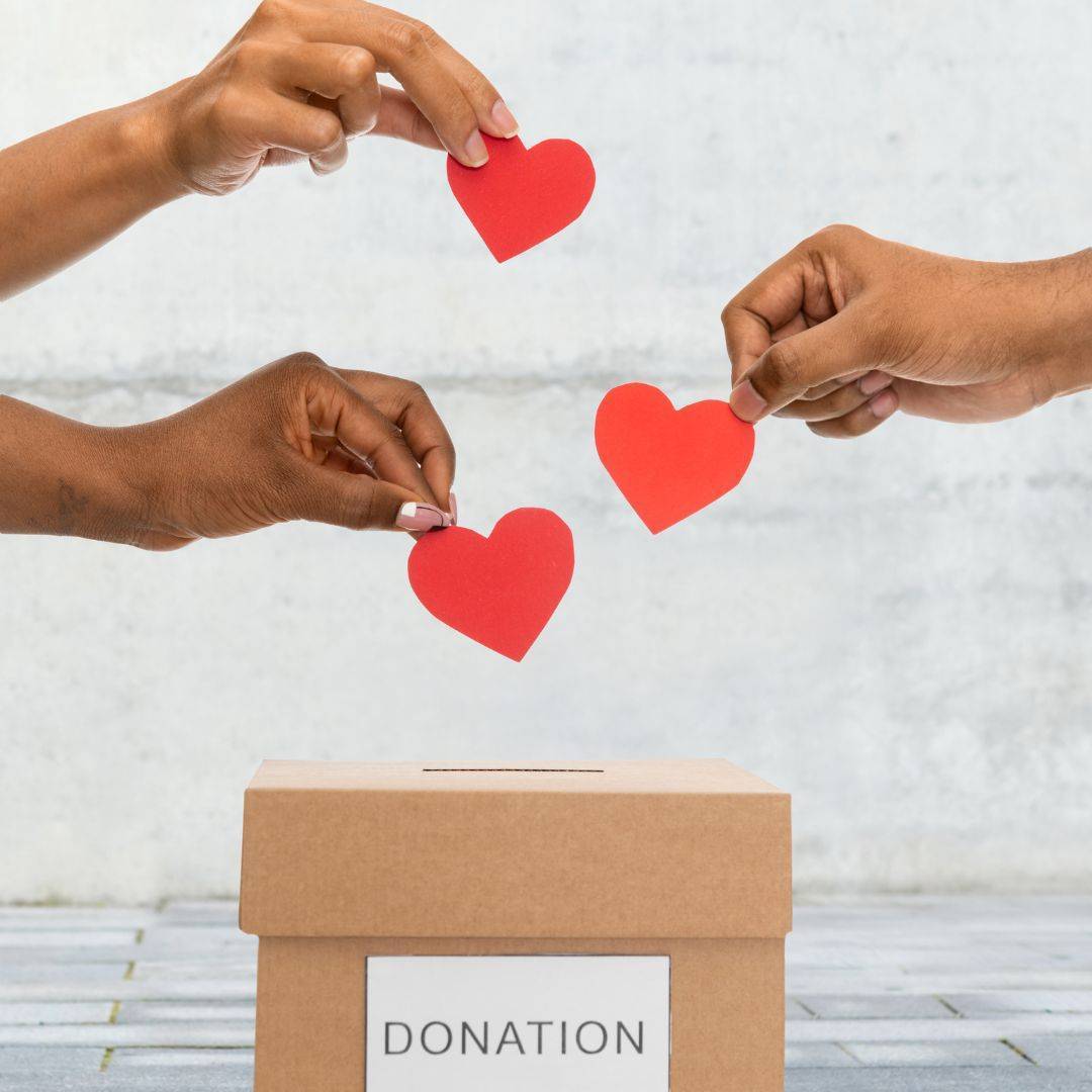 Hands placing red heart cutouts into a donation box. Symbolizing support and contributions towards a meaningful cause.