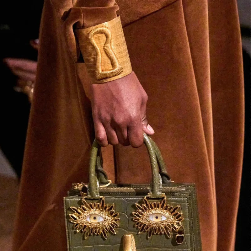 Gold exaggerated keyhole hair cuff styled on wrist with brown outfit and statement handbag.