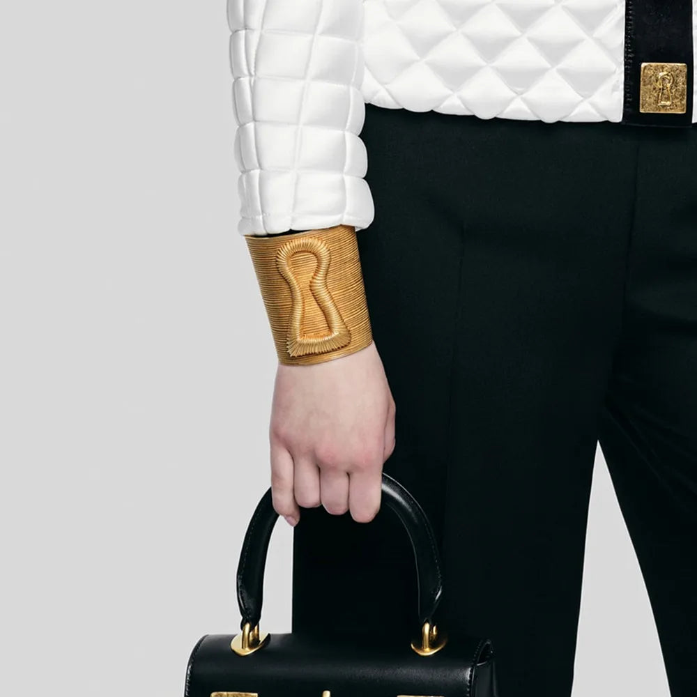 Gold keyhole hair cuff worn on wrist with white quilted jacket and black pants.