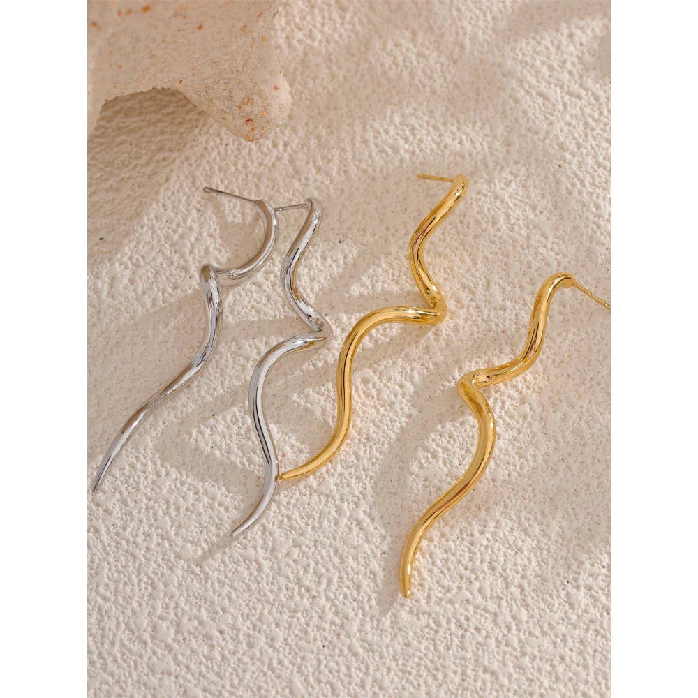 Gold and silver flowing metal geometric long stud earrings displayed on textured sand, showcasing their elegant wavy design.