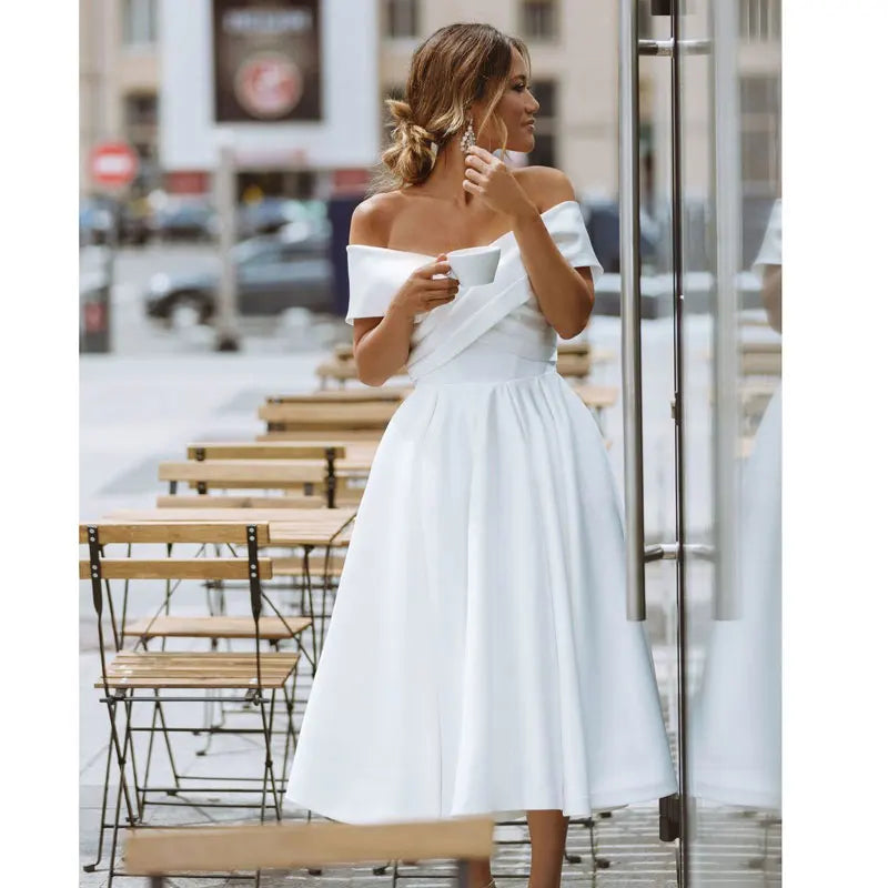 Bride-to-be in an off-shoulder white midi dress, standing outside a café with a coffee cup. A chic and modern wedding dress option.