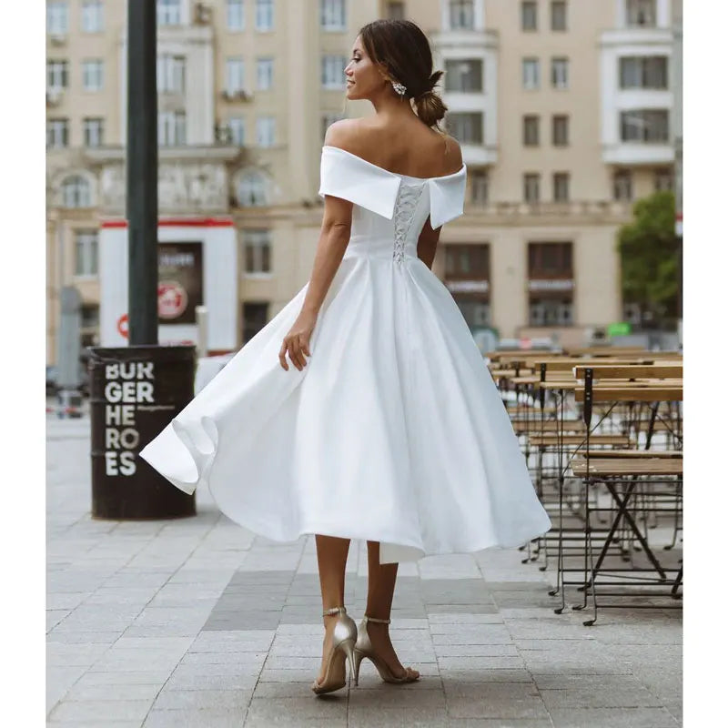 Back view of an off-shoulder white midi dress featuring a lace-up corset and flowing A-line skirt. Ideal for bridal and formal events.