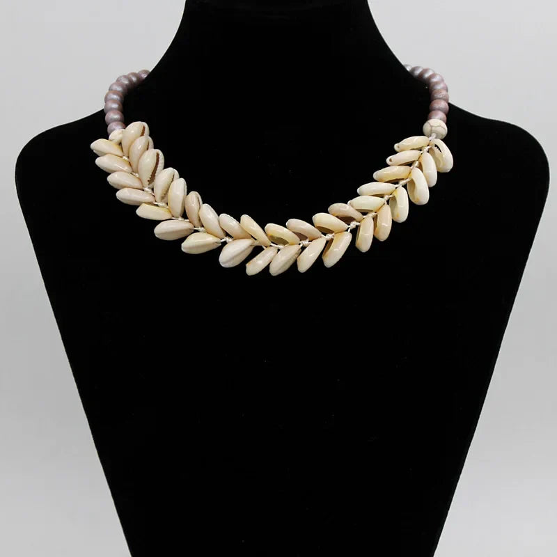 Handmade Cowrie Shell Necklace with purple beads on a black velvet bust. A stylish, natural shell accessory perfect for casual or beachwear.