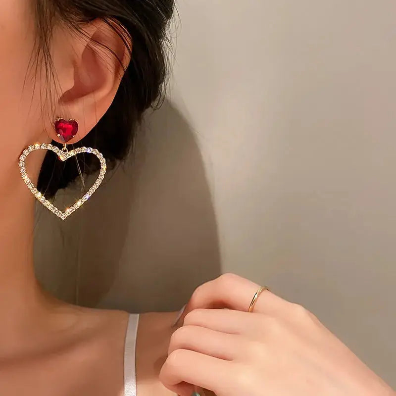 Model wearing heart-shaped crystal earrings with a red gemstone accent. A glamorous and elegant statement piece.