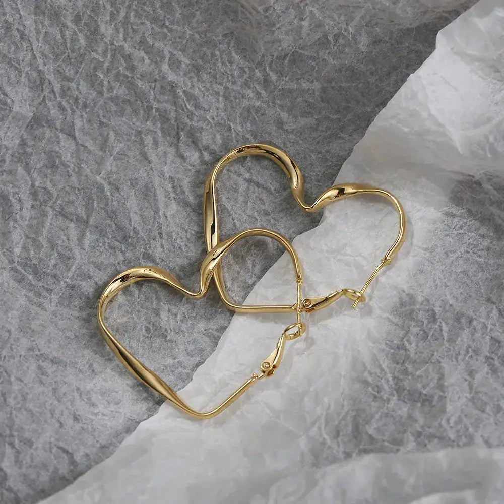 Gold heart-shaped hoop earrings with a wavy design, displayed on a textured surface. A chic and modern jewelry piece.