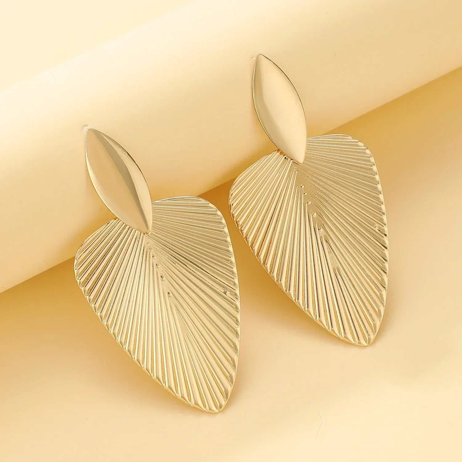 Gold fan statement earrings with a sleek and textured design, elegantly displayed on a soft yellow background. A perfect accessory for a bold style statement.