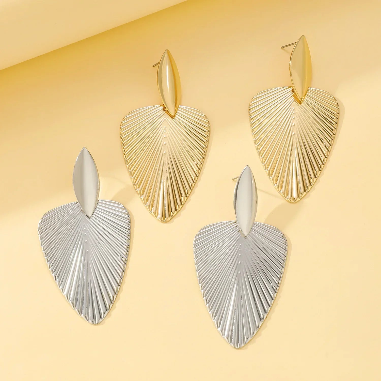 Gold and silver fan statement earrings with ribbed texture displayed on a soft yellow background. Stylish and elegant jewelry for a bold fashion statement.