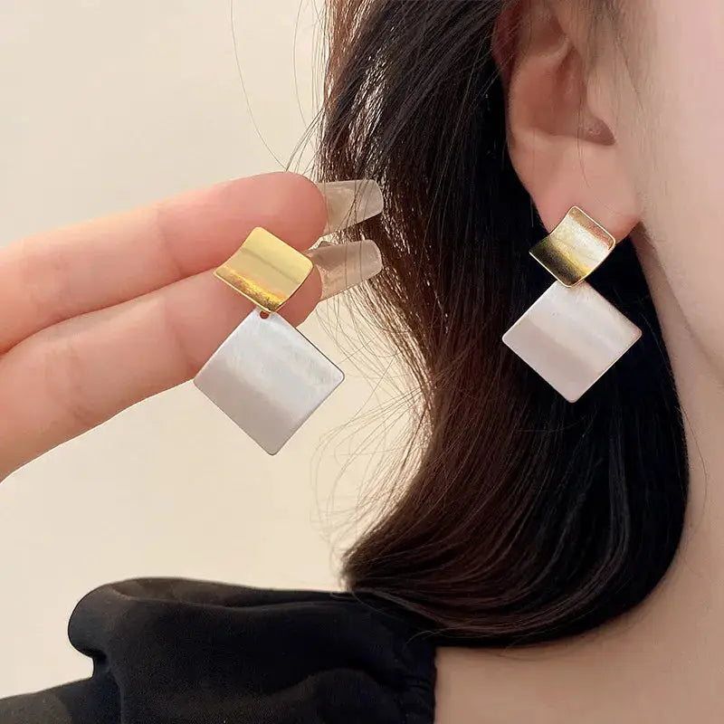 Geometric gold and white earrings with a modern square design, held by a model. A stylish and contemporary jewelry piece.