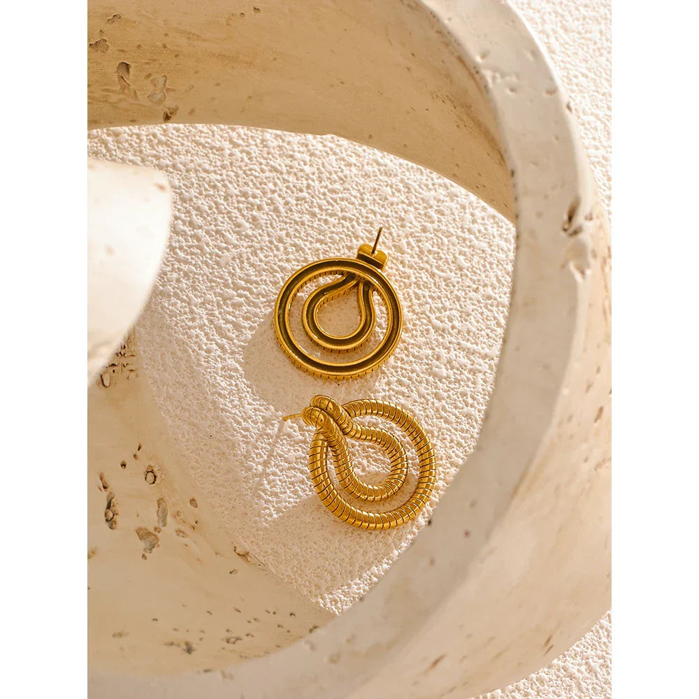 Close-up of stainless steel geometric snake earrings in gold-tone placed on a sculptural background. Elegant and unique jewelry piece.