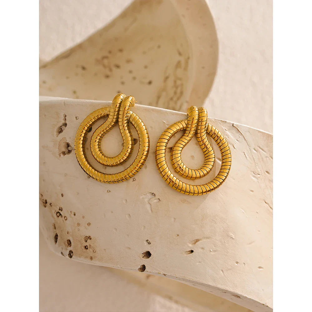 Gold-tone stainless steel geometric snake earrings resting on a curved stone surface. A vintage-inspired statement piece for women.