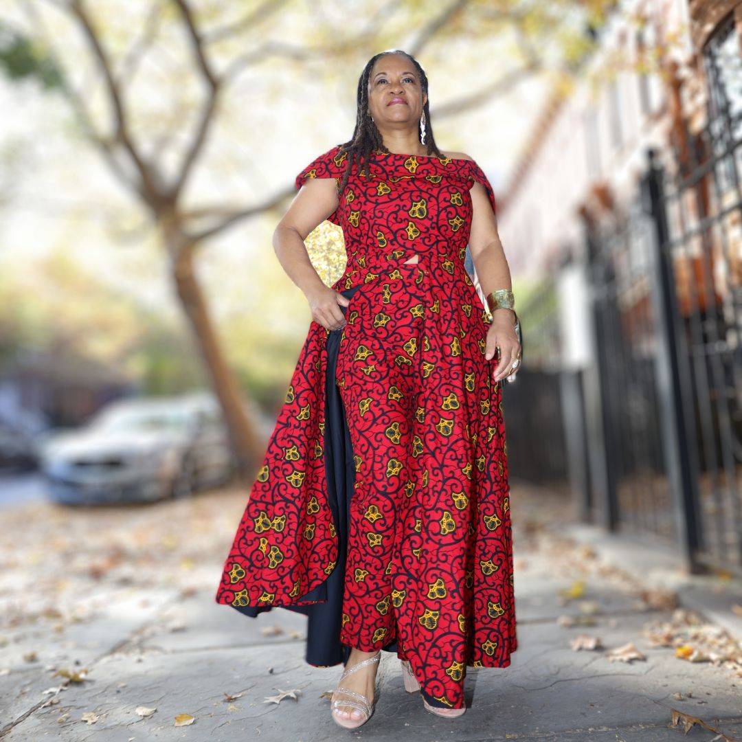 Confident woman in an African pantsuit with red and black print. Off-shoulder top with a dramatic cape, tailored for a custom fit in 100% cotton.