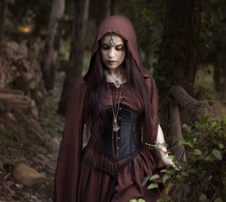 Woman in a brown hooded medieval maxi dress with a black corset, walking through a forest with an ethereal, gothic aesthetic.