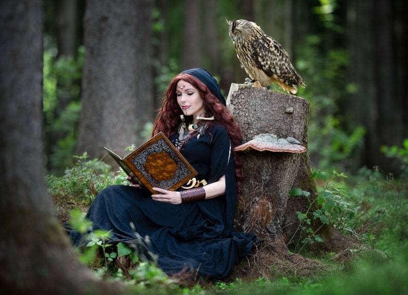 Boho-style hooded maxi dress in black, worn by a model reading an ornate book in a forest, paired with an owl for an enchanting look.