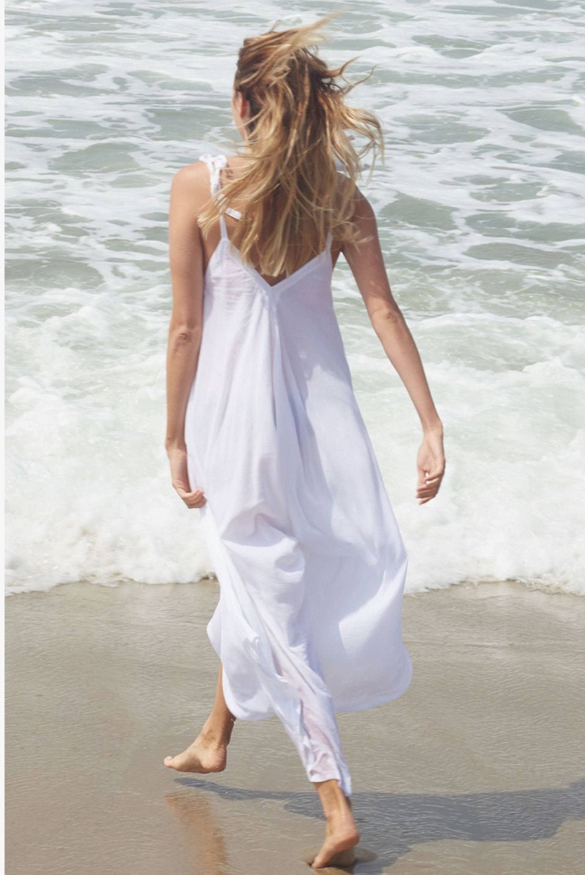 Woman walking along the beach in a white spaghetti angled straps jumpsuit with loose leg, highlighting its airy and lightweight fabric.