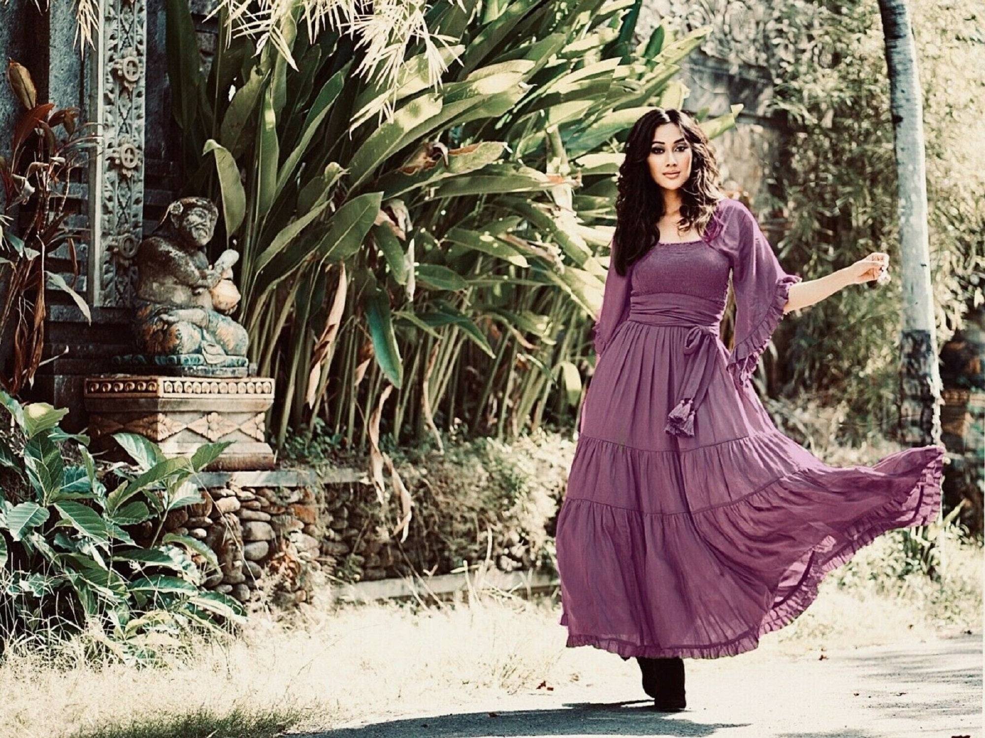 Purple medieval vintage maxi dress with an elasticized empire waist and tassel belt, flowing gracefully in a tropical garden. A dreamy boho outfit for all sizes.
