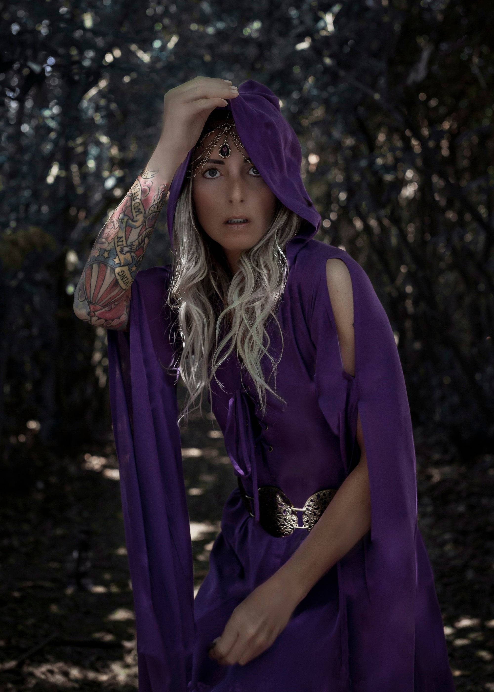 Mystical purple hooded medieval maxi dress with a gold belt, worn by a woman with a tattooed arm in a dark forest setting.