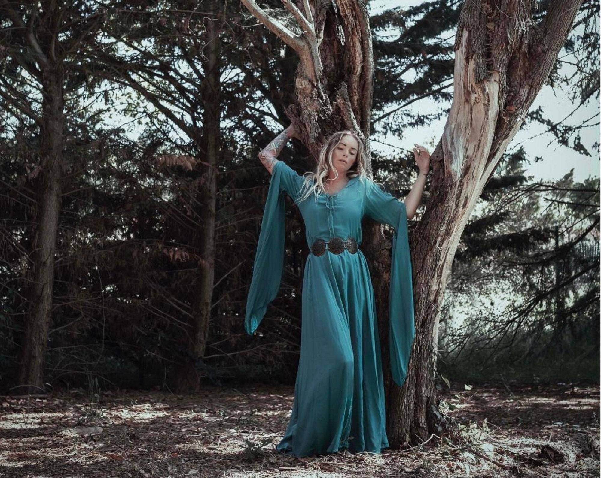 Bohemian-style medieval maxi dress in teal with flowing sleeves, worn by a woman leaning against a tree in a woodland setting.