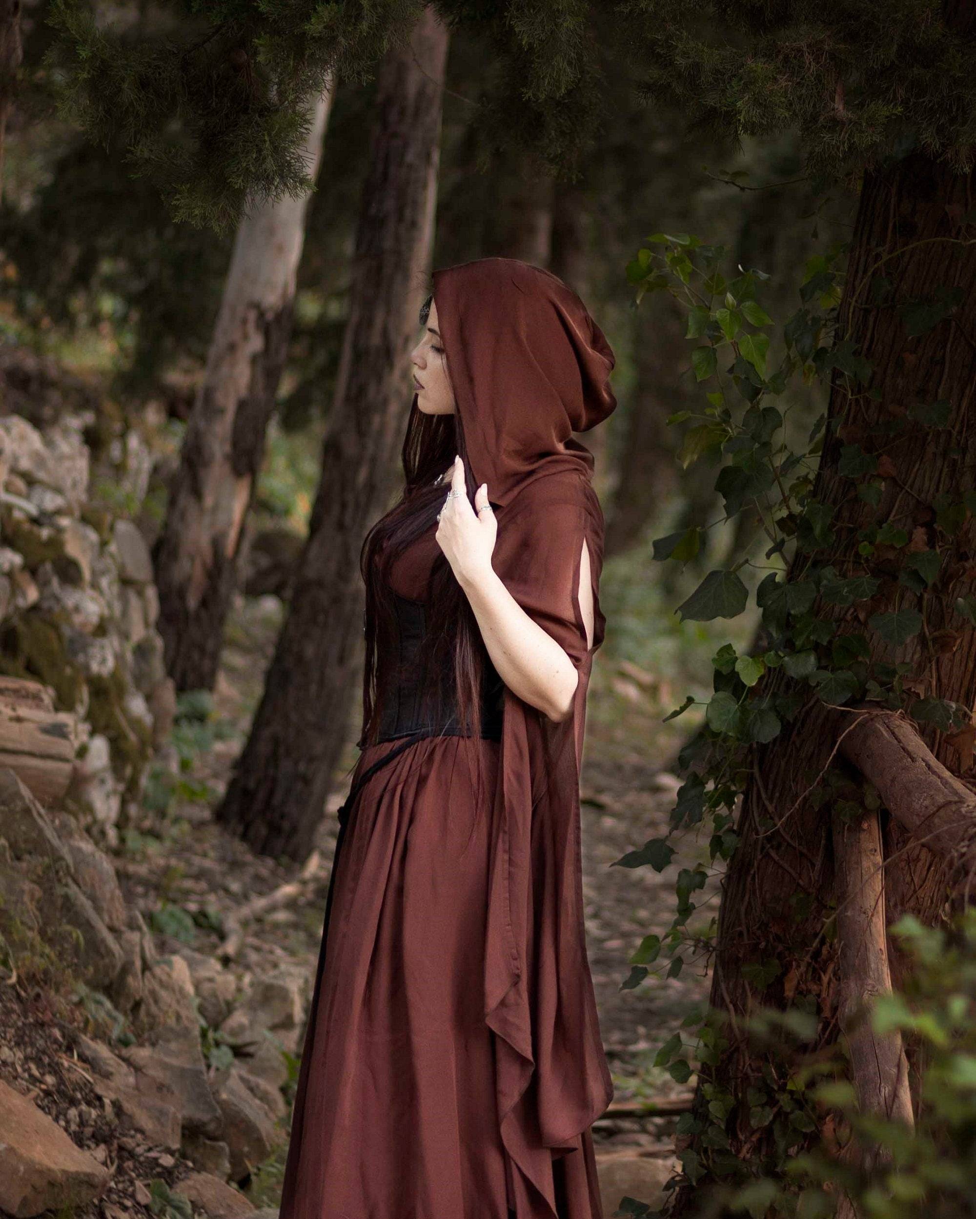 Side view of a woman adjusting the hood of her brown medieval maxi dress, highlighting its flowing fabric and fantasy appeal.