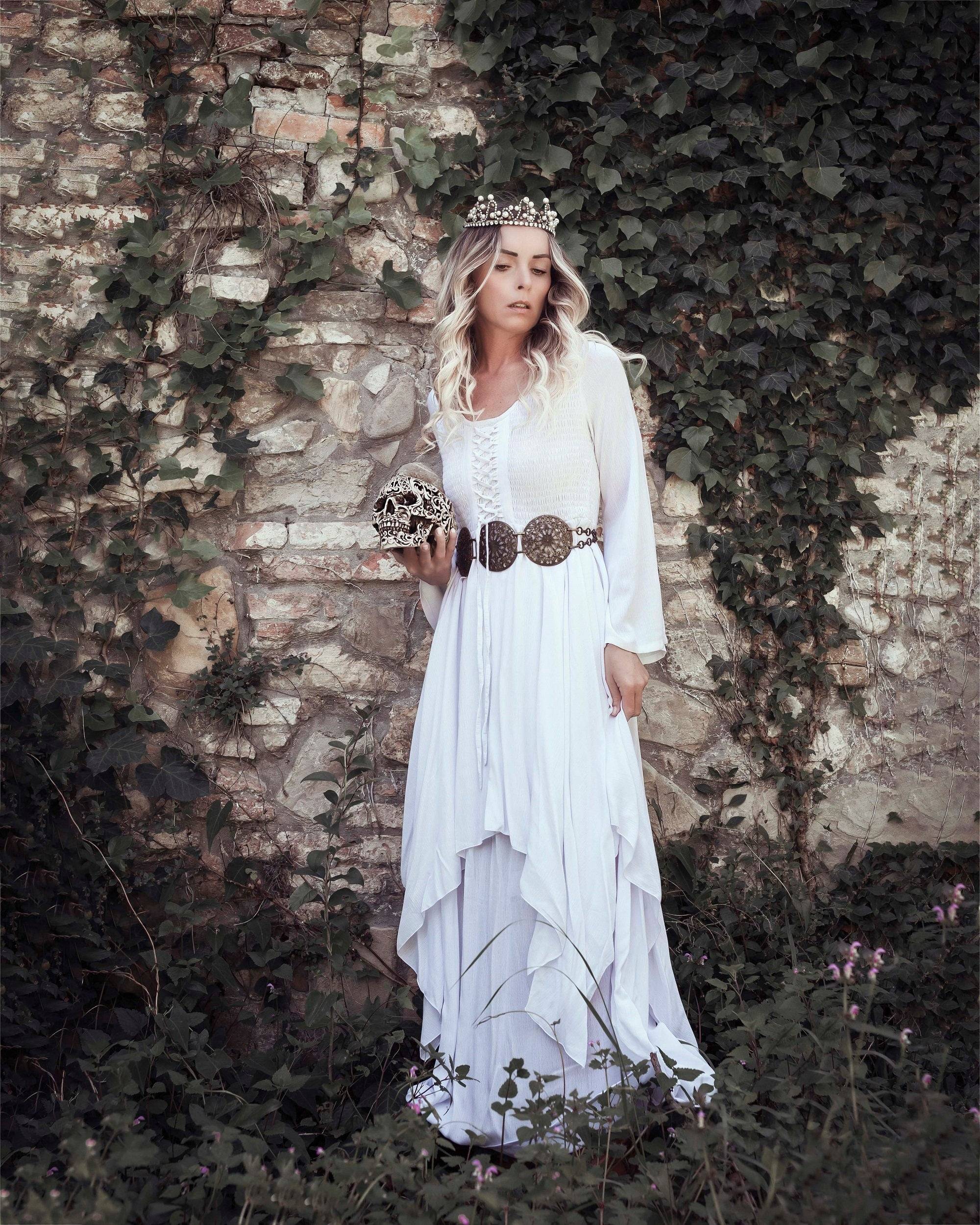 White Renaissance-style maxi dress with a lace-up bodice and layered skirt, accessorized with a statement belt and crown, set against a rustic stone wall.