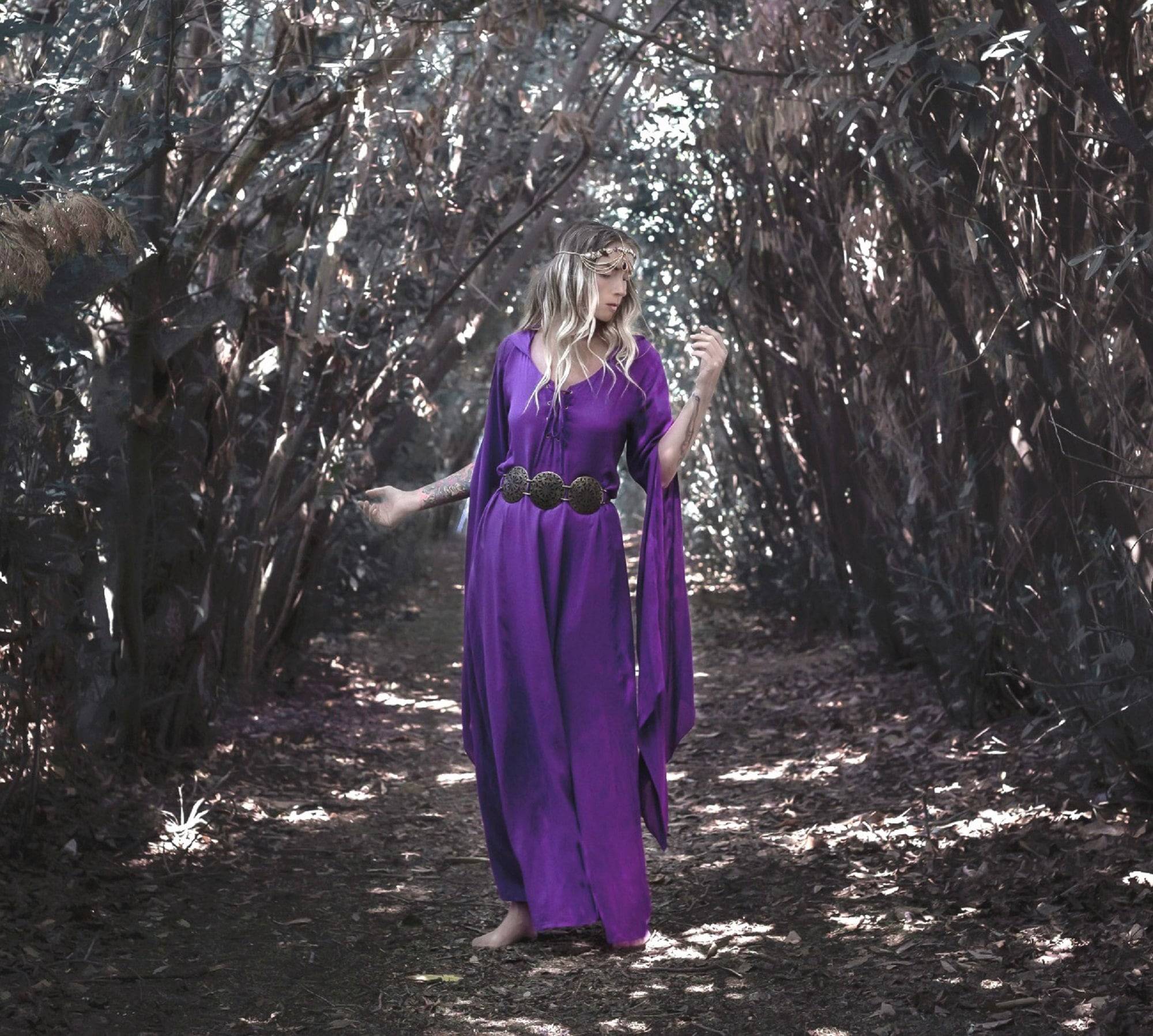 Fantasy-inspired purple medieval maxi dress with a hood and flowing sleeves, worn by a woman walking barefoot through a forest path.