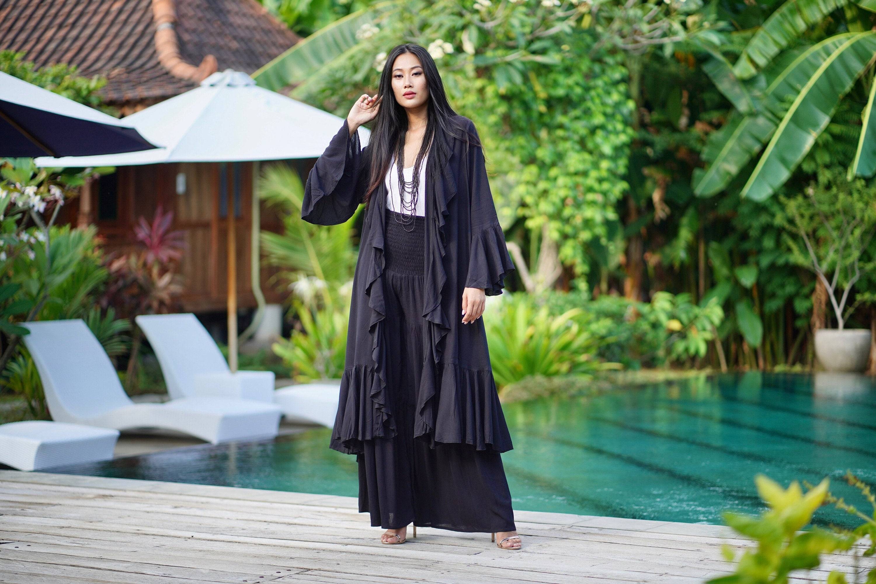 Black kimono-style wrap dress with ruffle details and a wide belt, styled elegantly by a poolside with tropical greenery.