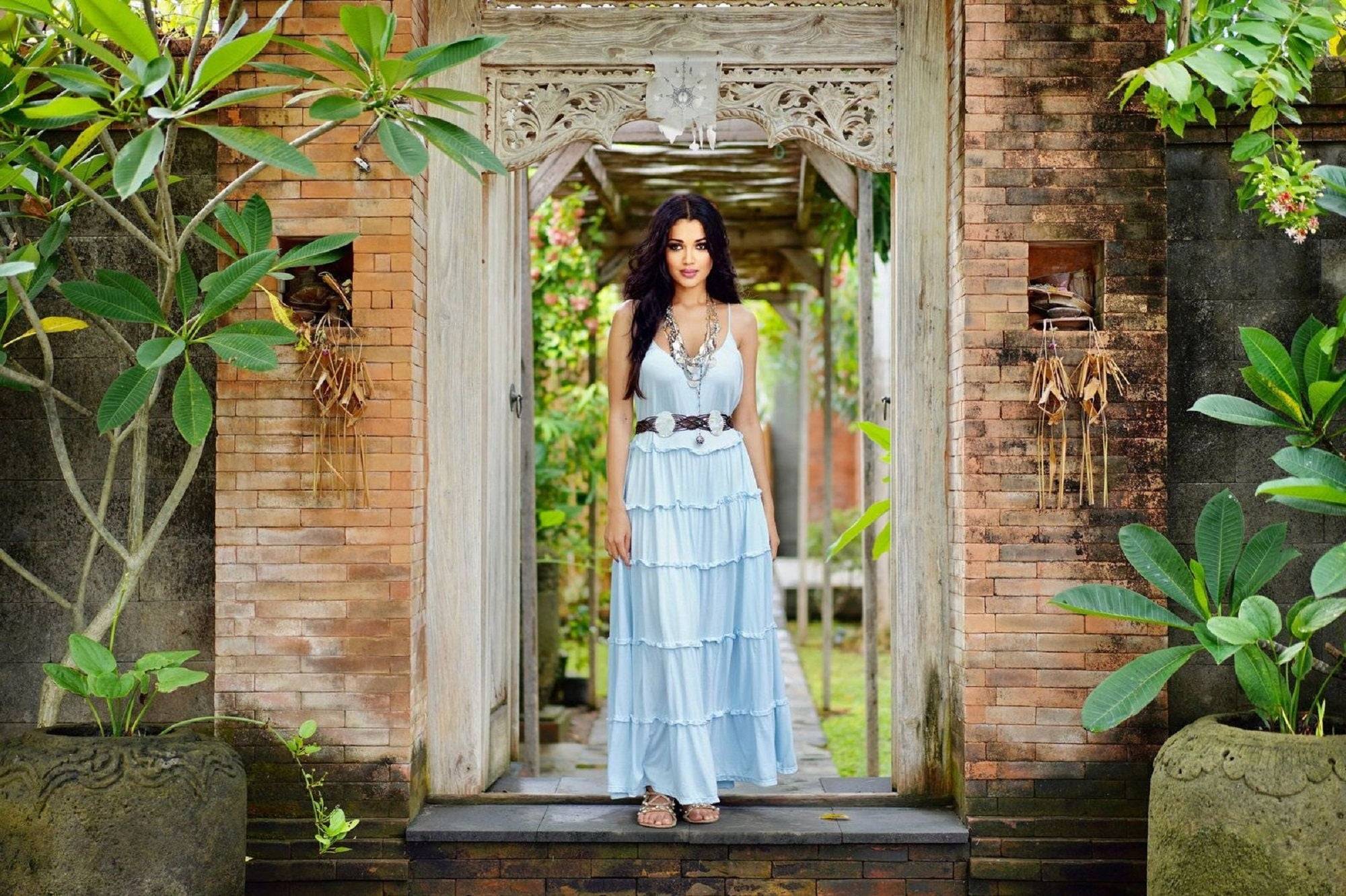 Light blue full-length stretchable jersey maxi dress with tiered ruffles, worn by a model in a tropical garden. Boho-chic fashion for all sizes.