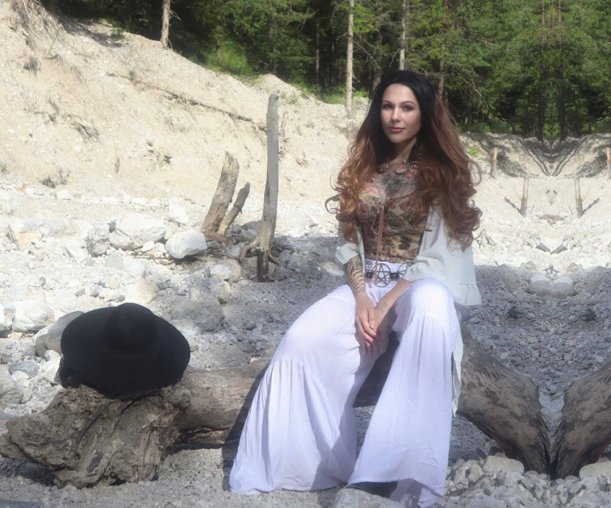 Relaxed bohemian look featuring white palazzo ruffled pants with smocked waist, styled with a floral top and black hat on a log.