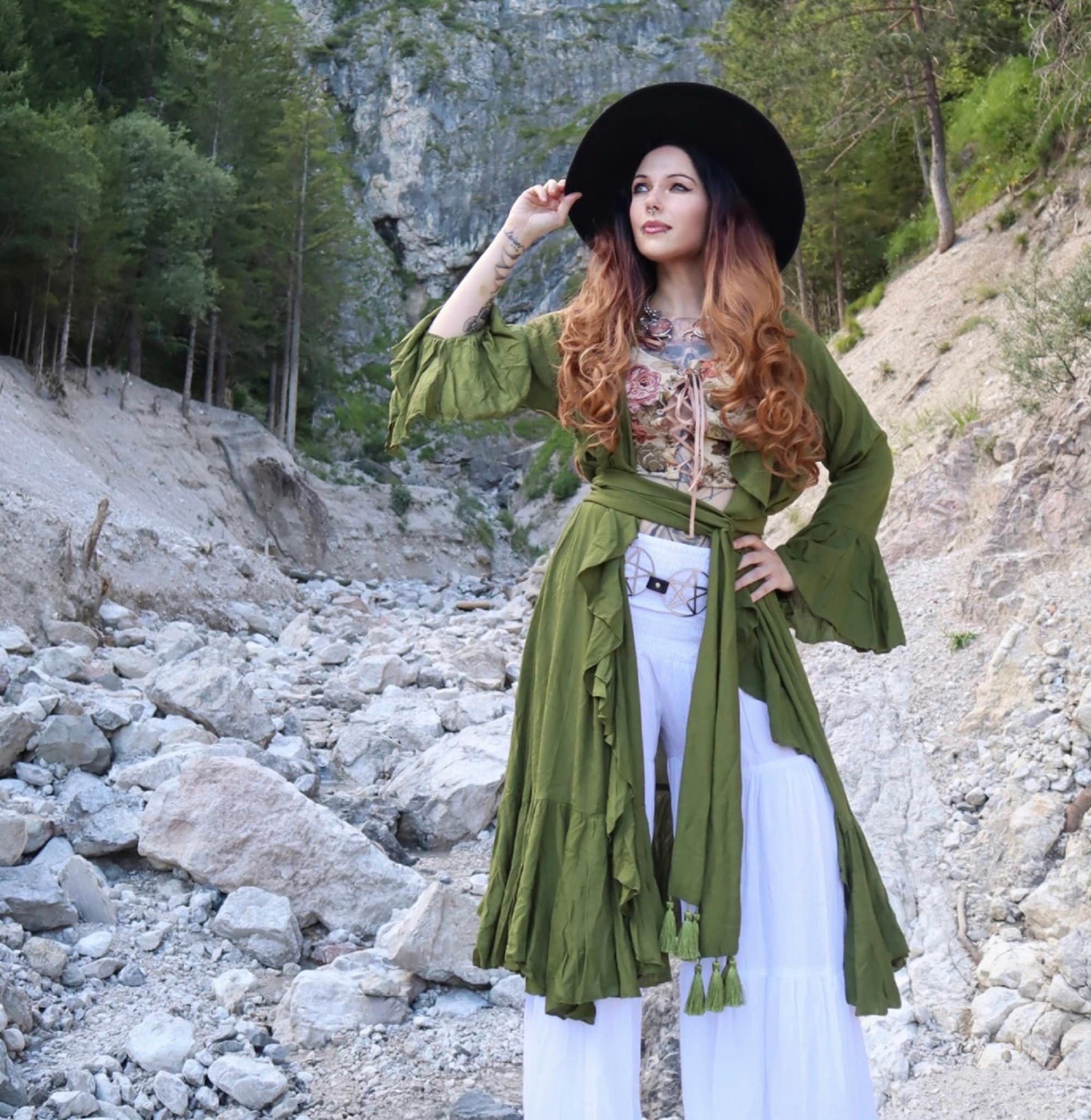 Stylish green kimono wrap dress with ruffle accents and a wide belt, worn with a black hat in a rocky landscape.