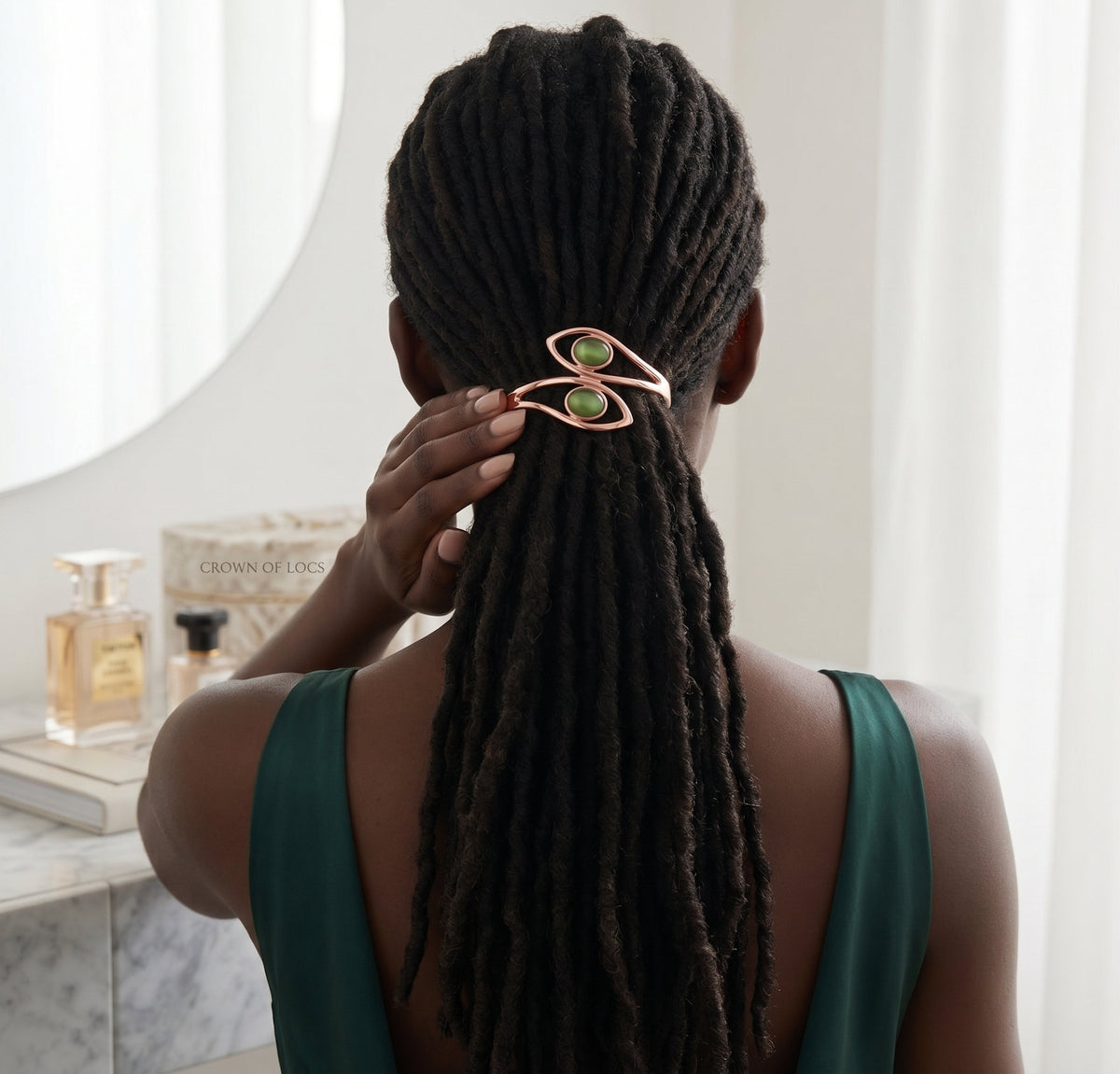 A person shown from behind with long dark dreadlocks secured by a rose gold hair claw clip with two green oval stones. The person is wearing a dark green top, standing in a bright indoor setting with a vanity and perfume bottles visible in the background.