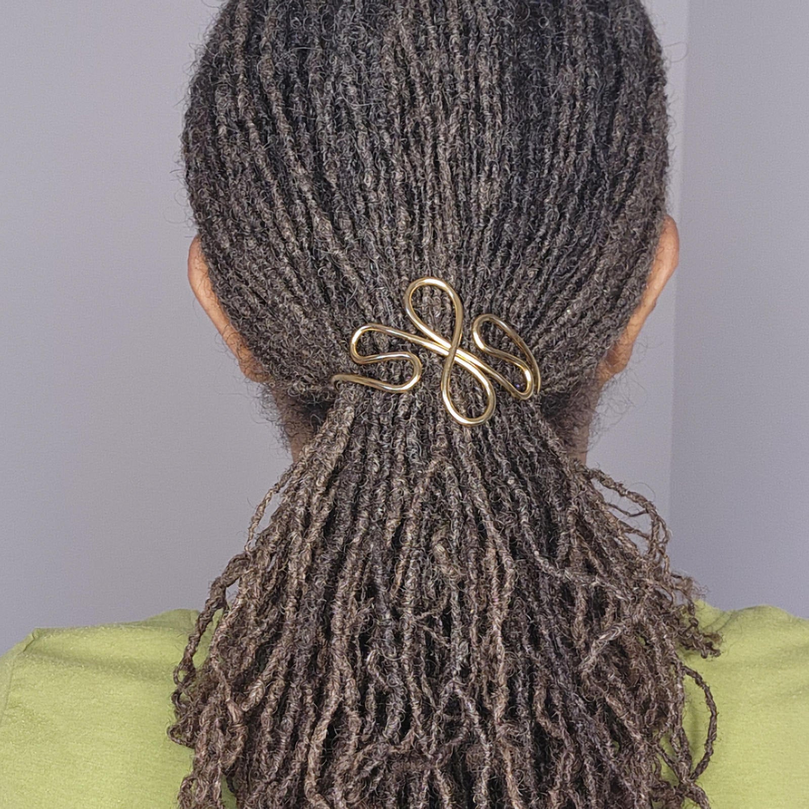 A close-up back view of a person with dark brown to black textured dreadlocks or a low bun secured with a shiny gold infinity-shaped hair claw clip. The person is wearing a light green top against a plain light gray background.