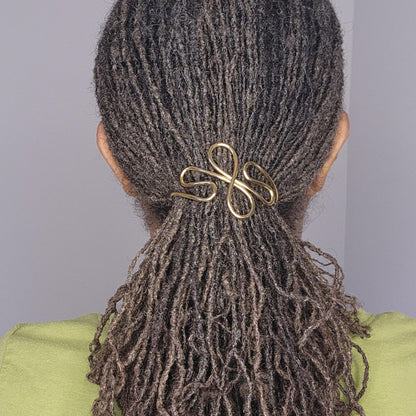 A close-up back view of a person with dark brown to black textured dreadlocks or a low bun secured with a shiny gold infinity-shaped hair claw clip. The person is wearing a light green top against a plain light gray background.