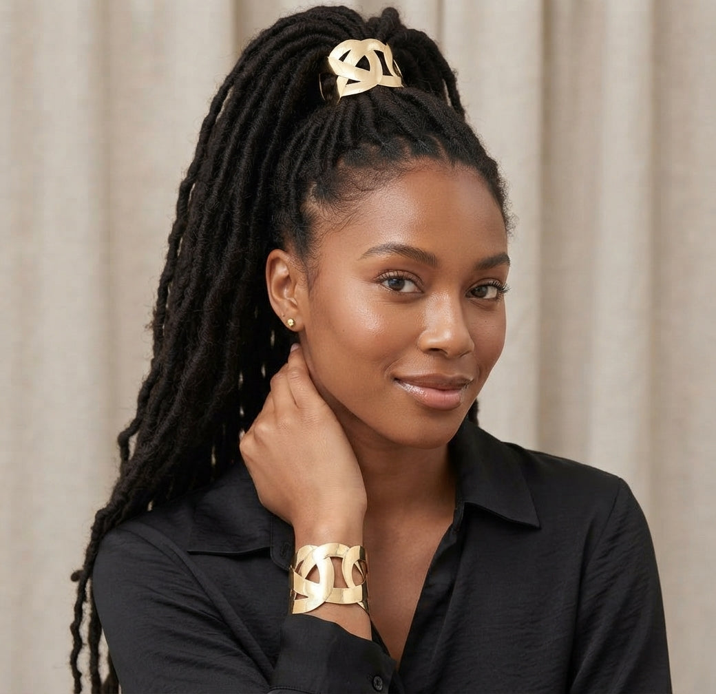Smiling woman with locs in a high ponytail, wearing a gold interlocking-loop hair cuff and a matching cuff on her wrist, in a black collared shirt.