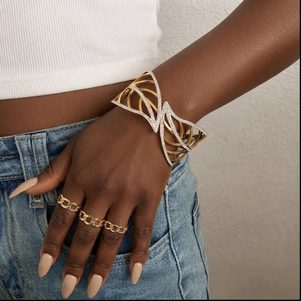 Wrist wearing a gold leaf-shaped openwork cuff with crystal edging, styled with jeans, a white crop top and gold chain rings