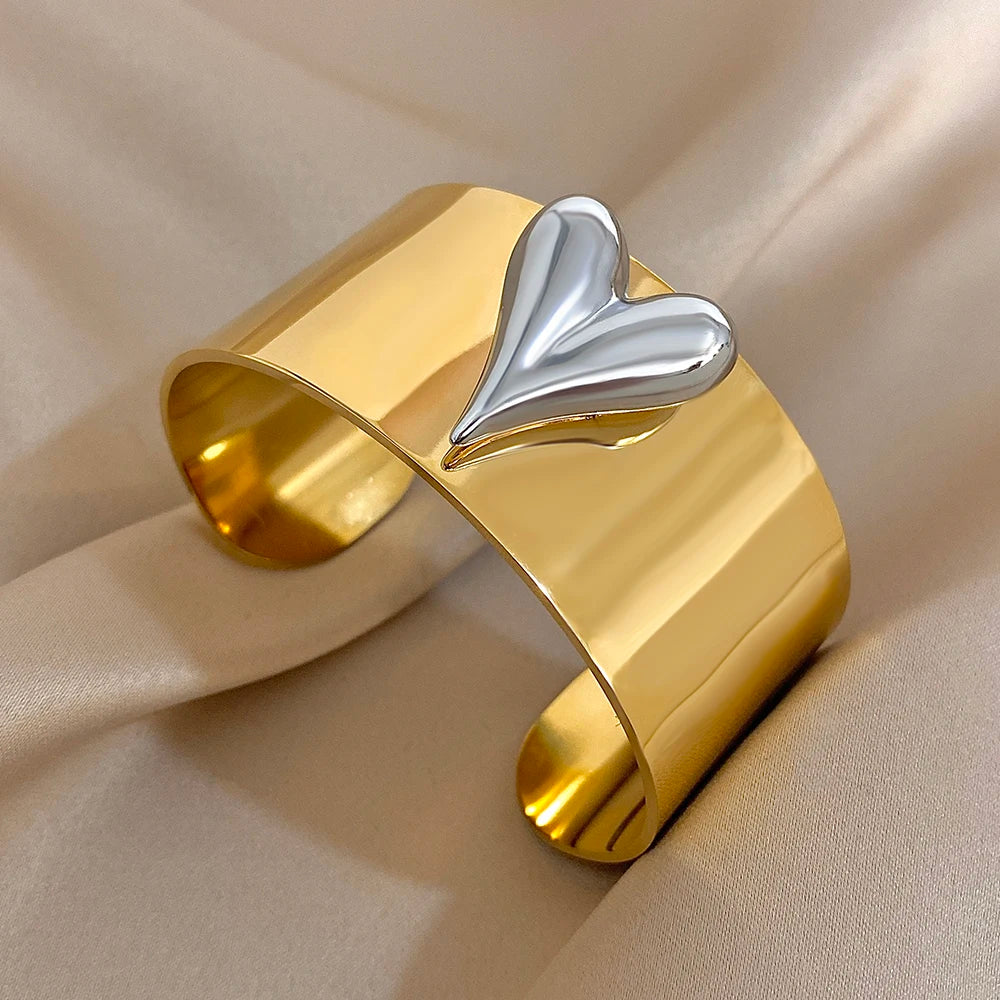 Gold stainless steel cuff bracelet featuring a raised silver puff-style heart on a polished wide band.