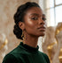 A woman with her hair in a low bun wears long, elegant gold dangle earrings with a wavy, snake-like design. She is wearing a dark green velvet turtleneck sweater, photographed in a warm, softly lit indoor setting with blurred decorative sculptures in the background.