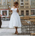 Woman in a white dress standing on a city street with tables and chairs in the background.