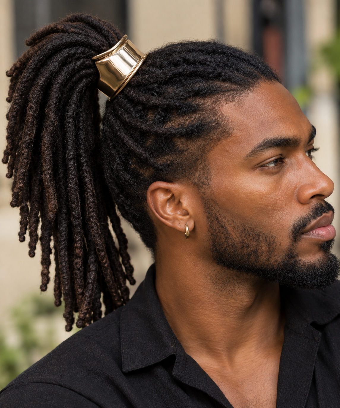 A close-up side profile of a Black man with long black dreadlocks tied back in a ponytail with a wide, chunky gold hair cuff for Locs. He is wearing a black collared shirt and a small hoop earring, set against a softly blurred outdoor background.