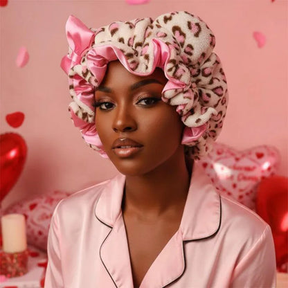 A woman wearing a plush leopard-print hair bonnet with pink satin ruffles and a matching pink satin pajama top, posed against a pink background with heart-shaped balloons.