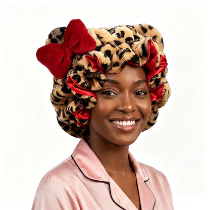 A smiling woman wearing a leopard-print hair bonnet with red velvet bows, paired with a light pink satin pajama top, set against a plain white background.