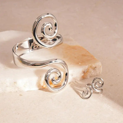 Sleek silver spiral cuff lying open on a neutral stone slab alongside a small matching silver spiral ring
