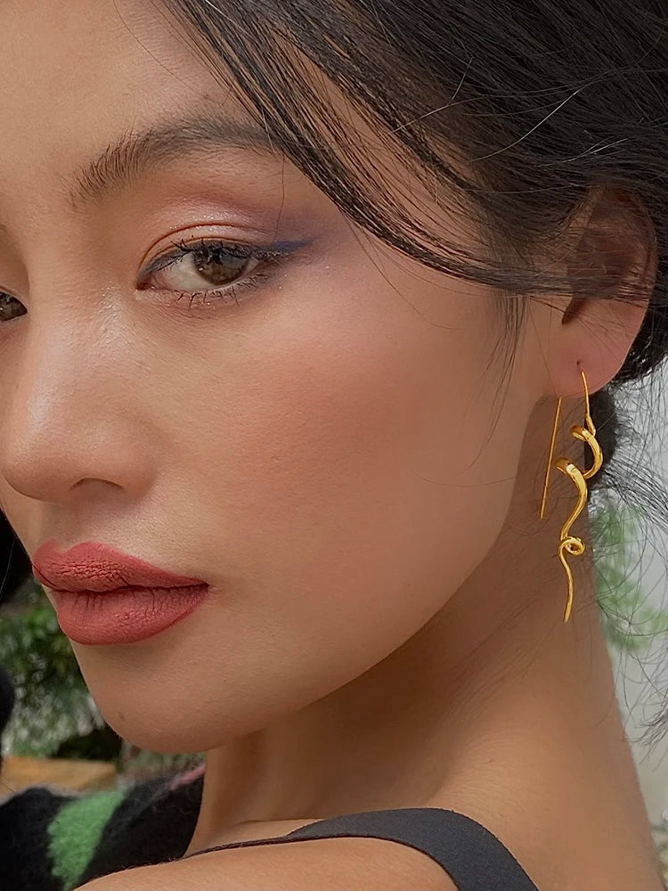 A close-up of a woman wearing a single gold dangle earring with a flowing, wavy spiral design. The shot focuses on her ear, jawline, and the jewelry, with soft natural light highlighting her skin and the metallic gold of the earring.