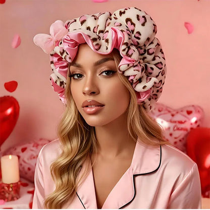 A woman wearing a plush leopard-print and pink satin hair wrap bonnet, paired with a matching pink satin pajama top, set against a romantic pink background with heart balloons and petals.