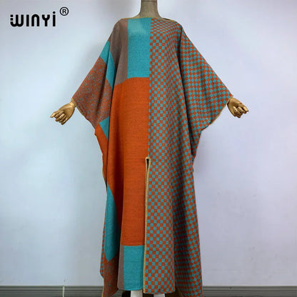 A long, loose woven poncho or caftan displayed on a mannequin against a plain light gray wall. The garment has a color-blocked design with solid orange, teal, and taupe sections, plus a small-scale check pattern in orange and teal on taupe.