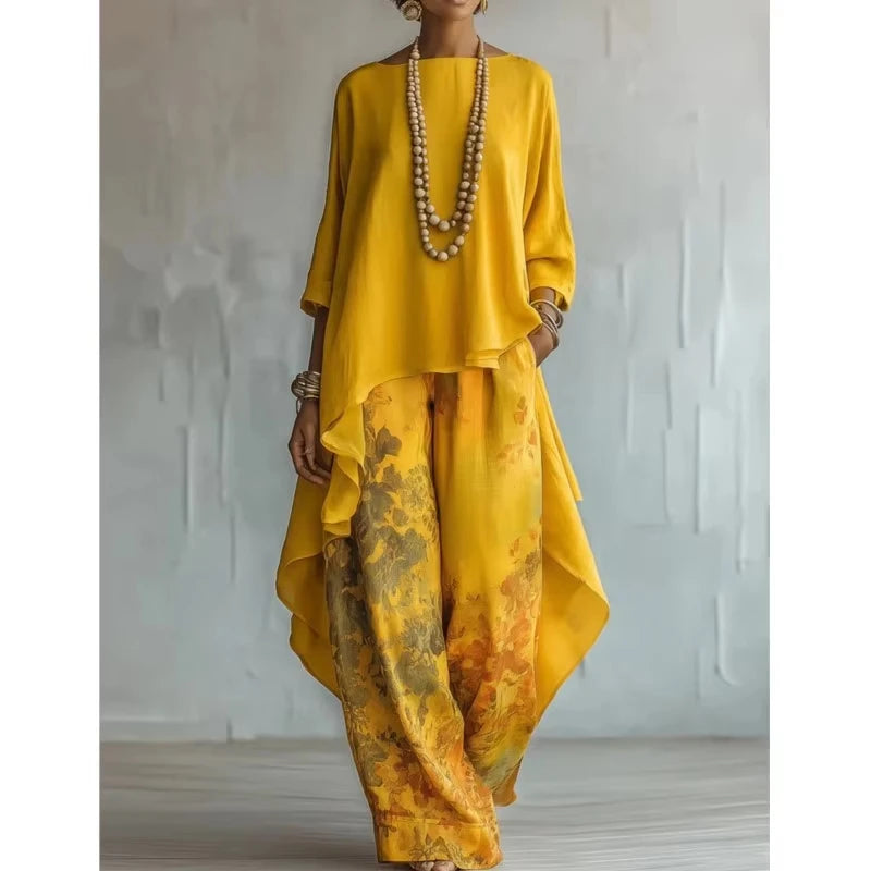 A woman models a two-piece outfit featuring a bright yellow high-low top and matching wide-leg pants with a yellow, orange, and brown abstract watercolor print. She is accessorized with a long beaded necklace and bracelets, standing against a textured light gray wall.