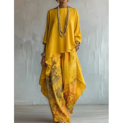 A woman models a two-piece outfit featuring a bright yellow high-low top and matching wide-leg pants with a yellow, orange, and brown abstract watercolor print. She is accessorized with a long beaded necklace and bracelets, standing against a textured light gray wall.