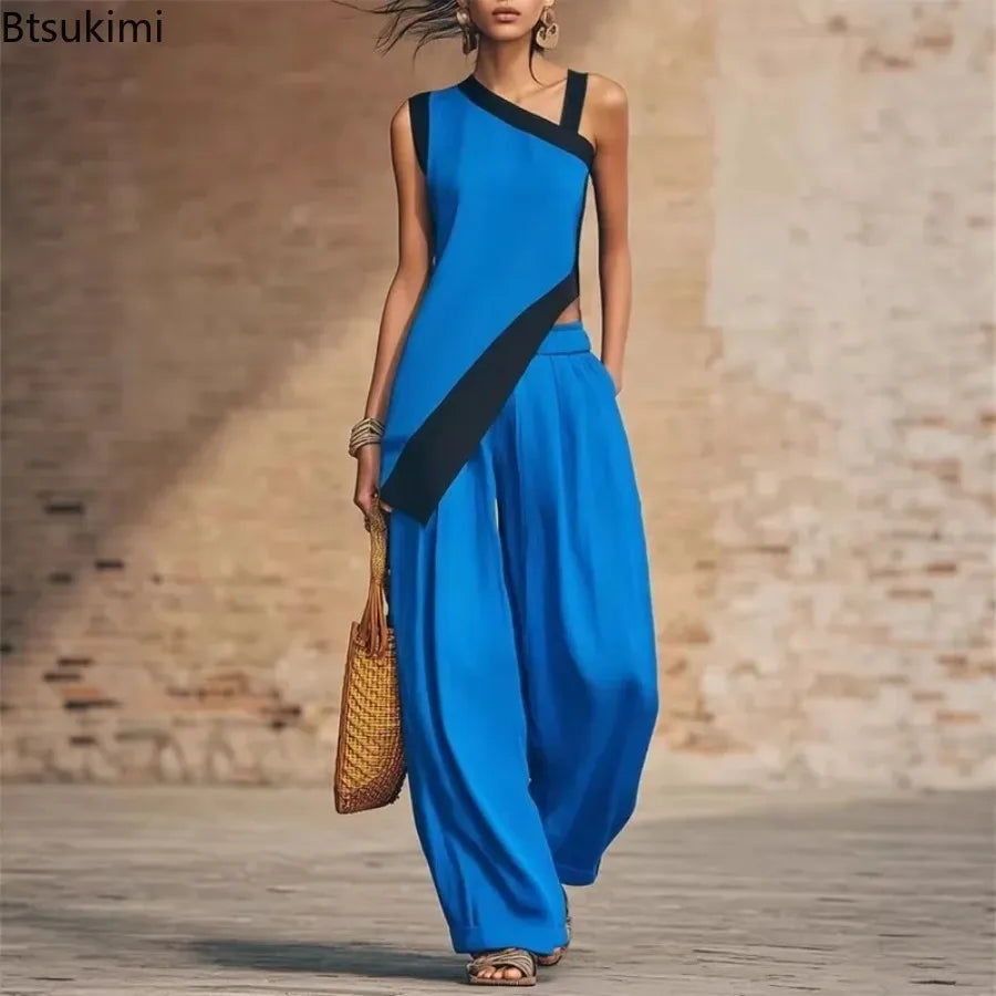 A woman models a vibrant blue two-piece outfit consisting of an asymmetrical one-shoulder top and matching wide-leg pants, styled with a woven straw bag against a rustic stone wall background.