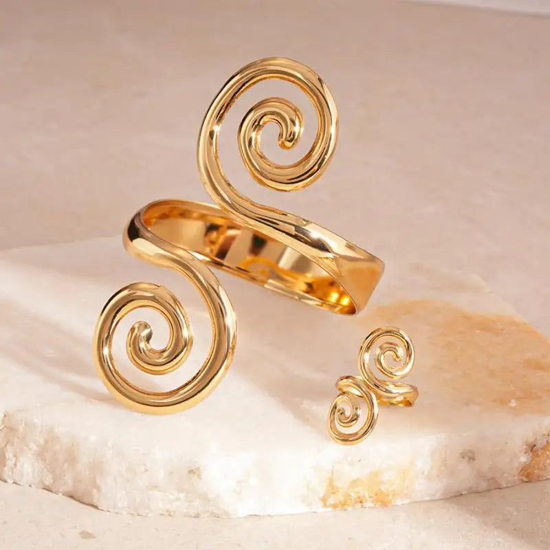 High-shine gold spiral cuff standing vertically next to a mini gold spiral ring on a light beige stone surface.