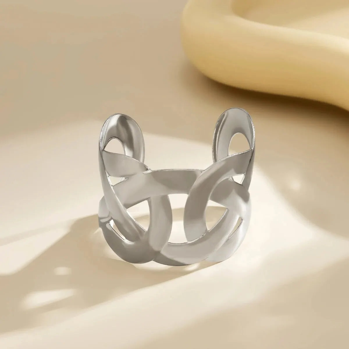 Silver open cuff with interlocking looped cutouts, standing on a cream surface with soft shadows.