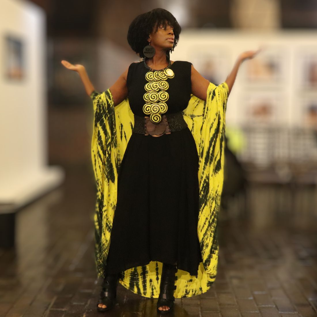 Stylish black maxi dress with a bold yellow tie-dye cape, accessorized with a statement belt and jewelry. Ideal for a chic, artistic look.